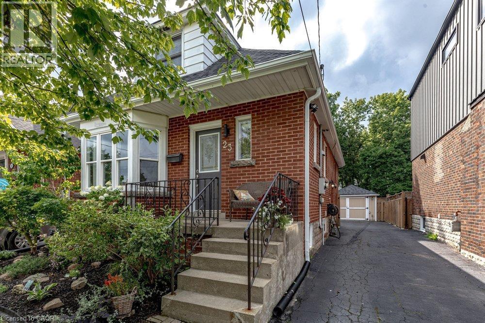 Brick exterior with white trim, front entrance with concrete steps and wrought iron railing, bay window, mature front landscaping, and a paved driveway leading to a detached garage - 23 Kipling Road, Hamilton, ON - Outdoor With Exterior