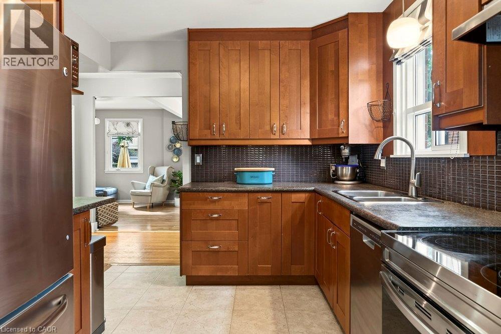 Kitchen featuring wood cabinetry, stainless steel appliances, dark countertops, a dark tile backsplash, and light tile flooring - 23 Kipling Road, Hamilton, ON - Indoor Photo Showing Kitchen With Double Sink