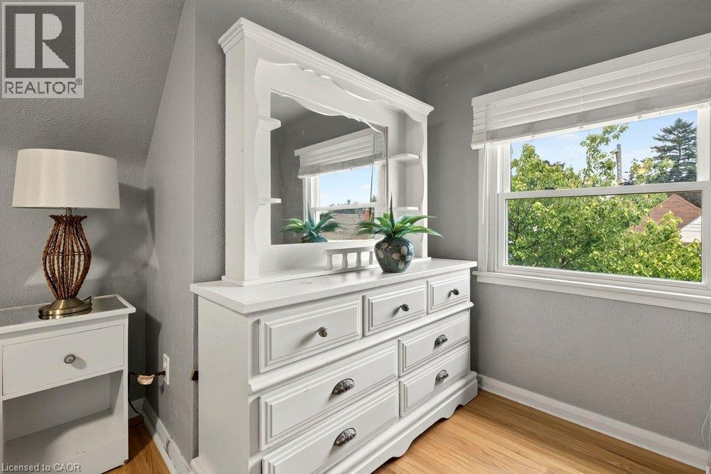 Bright secondary bedroom featuring a white dresser with mirror and multiple drawers, hardwood flooring, large windows with natural light, and gray wall finishes - 23 Kipling Road, Hamilton, ON - Indoor