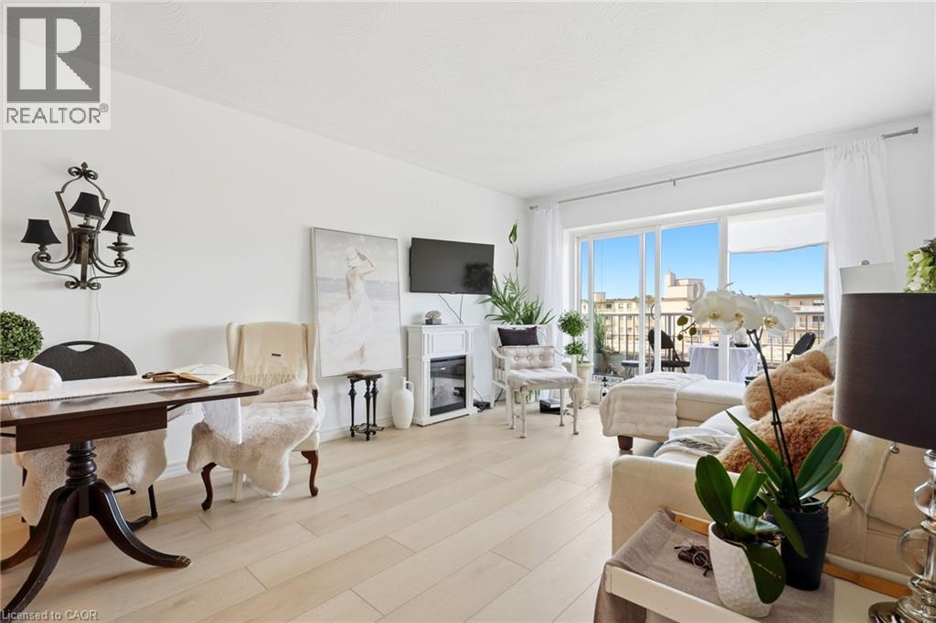 Light wood-finish flooring throughout, sliding glass doors to exterior, wall-mounted chandelier fixture, and built-in electric fireplace - 8 Woodman Drive S Unit# 708, Hamilton, ON - Indoor
