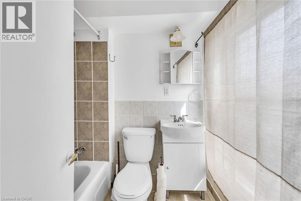 White tub with brown wall tile surround, white toilet, pedestal sink with vanity, medicine cabinet with mirror, and wall-mounted light fixture - 1367 Cannon Street E, Hamilton, ON - Indoor Photo Showing Bathroom