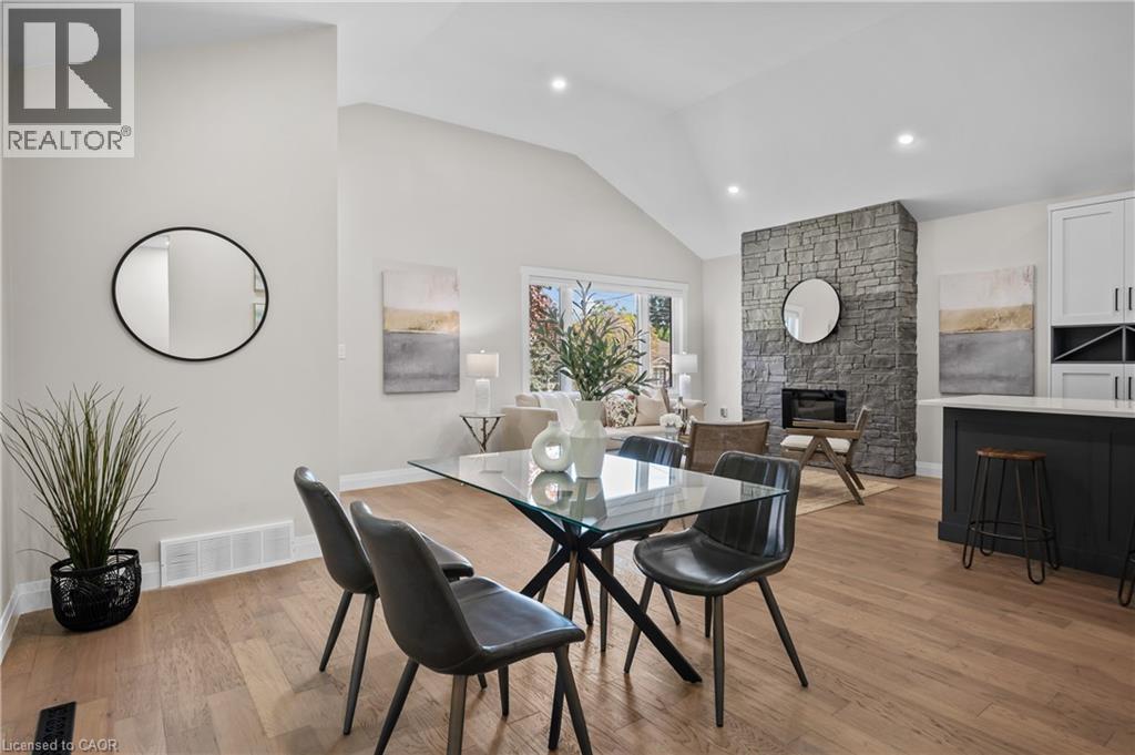 Open-concept dining and living area featuring a wood-finish plank floor, black metal dining table with seating for four, vaulted ceiling with recessed lighting, and a striking stone fireplace accent w - 1041 Shepherd'S Drive, Burlington, ON - Indoor Photo Showing Dining Room
