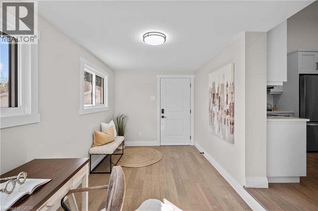 Wood-finish flooring, white walls, multiple windows, a white entry door, and a partially visible kitchen with white cabinetry - 1041 Shepherd'S Drive, Burlington, ON - Indoor