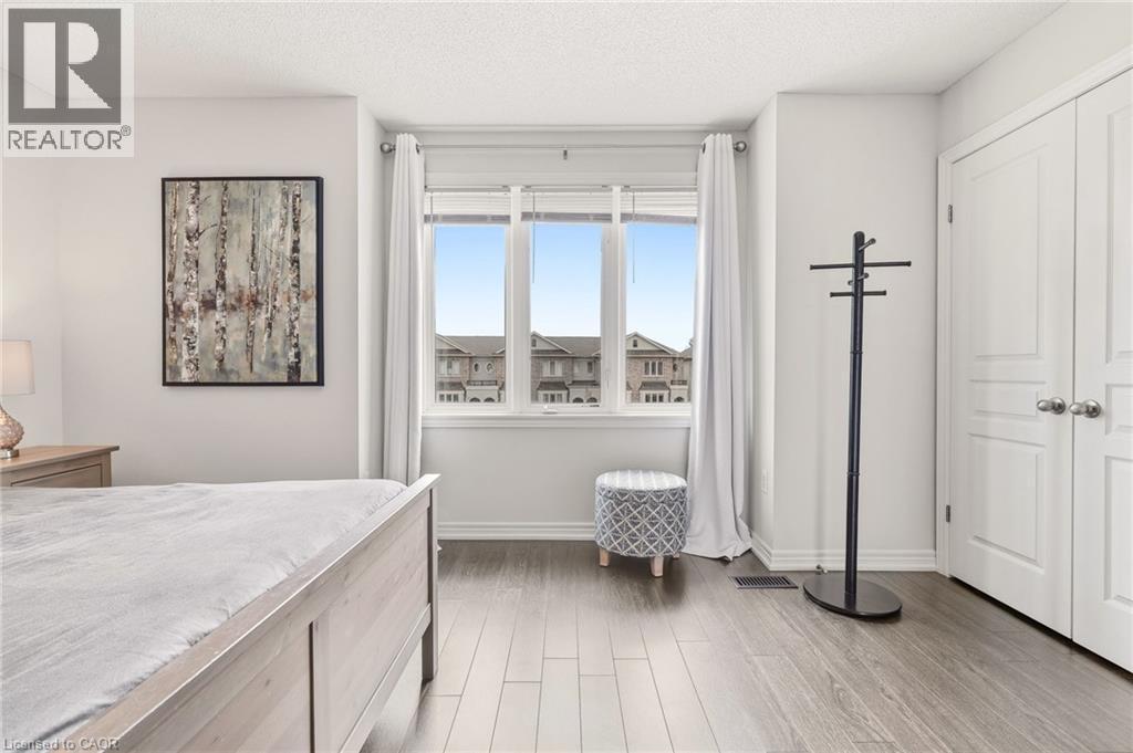 Bright primary bedroom featuring expansive windows with panoramic vistas, wood-finish flooring throughout, marble-topped bed frame, and direct access to a walk-in closet - 4171 Palermo Common, Burlington, ON - Indoor Photo Showing Bedroom
