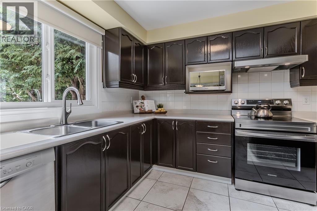 Dark wood cabinetry with brushed nickel hardware, white tiled flooring, stainless steel appliances, white subway tile backsplash, and a double basin sink with a gooseneck faucet - 1238 Sable Drive, Burlington, ON - Indoor Photo Showing Kitchen With Double Sink
