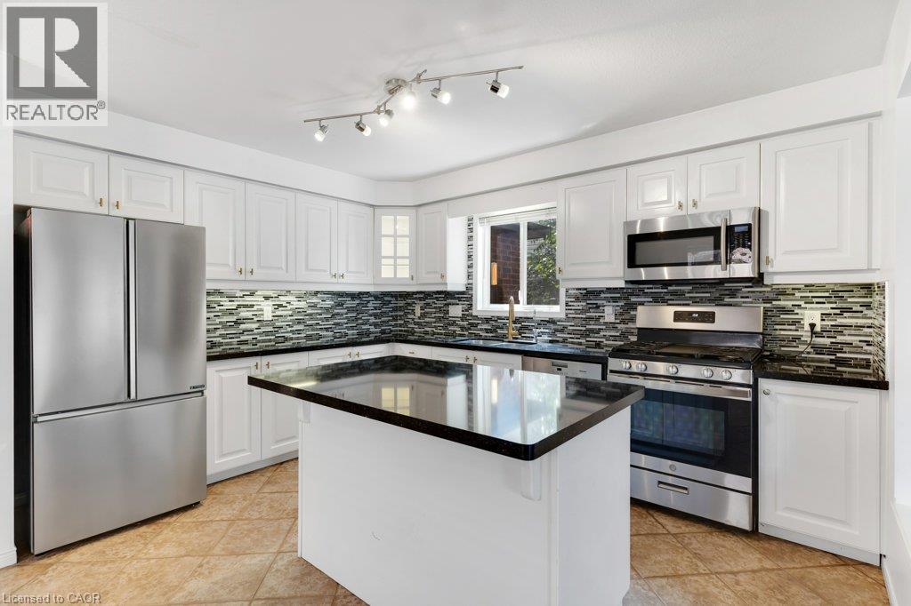 Kitchen featuring white cabinetry, a central island with a dark countertop, stainless steel appliances, a mosaic tile backsplash, and ceramic tile flooring - 76 Renner Drive, Cambridge, ON - Indoor Photo Showing Kitchen