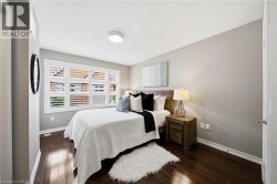 Bright secondary bedroom with wood-finish flooring, plantation shutters spanning the bay window, neutral wall finishes, and a wood nightstand with attached task lighting - 