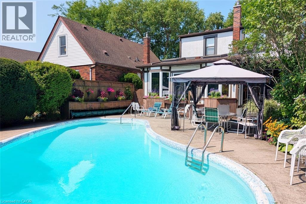 Curved swimming pool with concrete surround, mature landscaping, and an attached covered patio area - 107 Bond Street S, Hamilton, ON - Outdoor With In Ground Pool