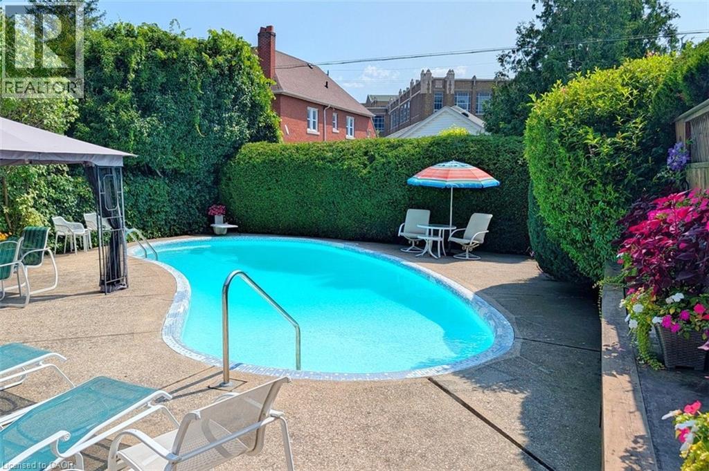 Freeform swimming pool with integrated steps, concrete pool deck, and dense perimeter hedging - 107 Bond Street S, Hamilton, ON - Outdoor With In Ground Pool With Backyard