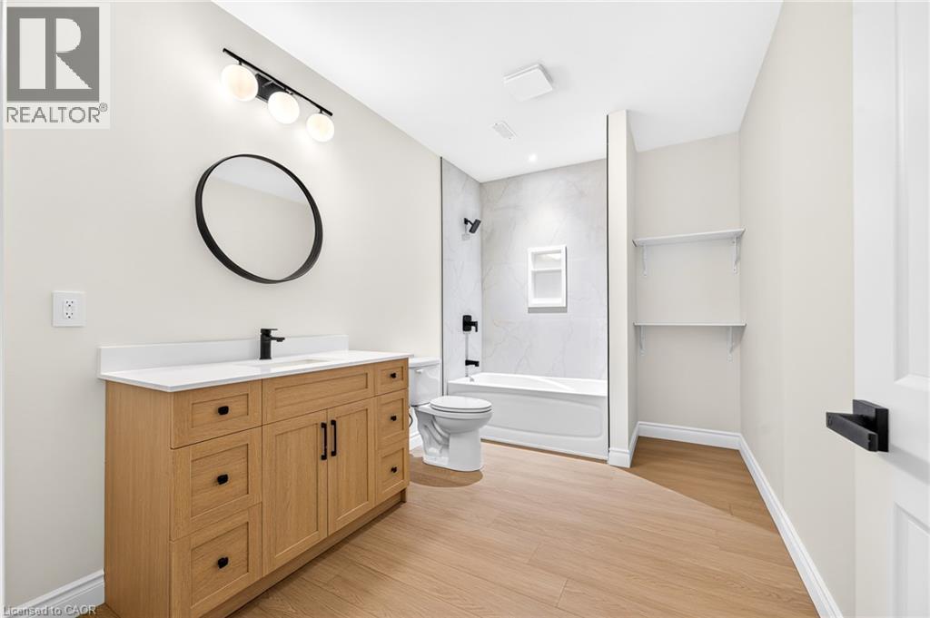 Wood-finish flooring, light wood vanity with white countertop, round wall mirror, and modern black fixtures - 67 Cielo Court, Hamilton, ON - Indoor Photo Showing Bathroom