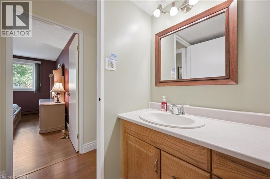 Wood-finish vanity with a white countertop and integrated sink, framed mirror, and three-light vanity fixture - 6 Walton Avenue Unit# 107, Kitchener, ON - Indoor Photo Showing Bathroom