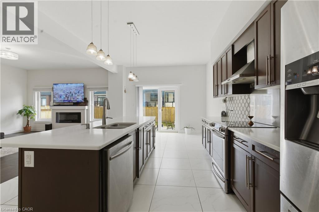 Galley-style kitchen featuring stainless steel appliances, white quartz countertops, dark wood cabinetry with glass-front upper cabinets, pendant lighting over the island, and tile flooring throughout - 2000 Maddex Way, London, ON - Indoor Photo Showing Kitchen With Double Sink With Upgraded Kitchen