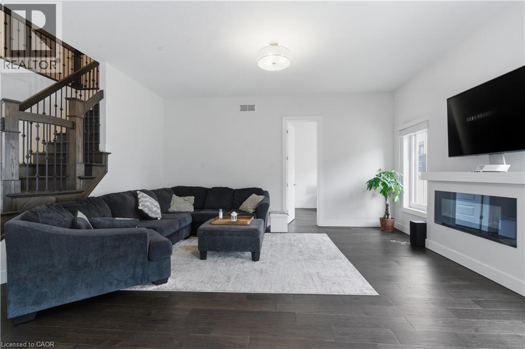 Spacious open-concept living area featuring dark wood-finish flooring, a curved sectional seating arrangement, a linear gas fireplace with white surround, wall-mounted flat-screen television, and abun - 2000 Maddex Way, London, ON - Indoor Photo Showing Living Room With Fireplace