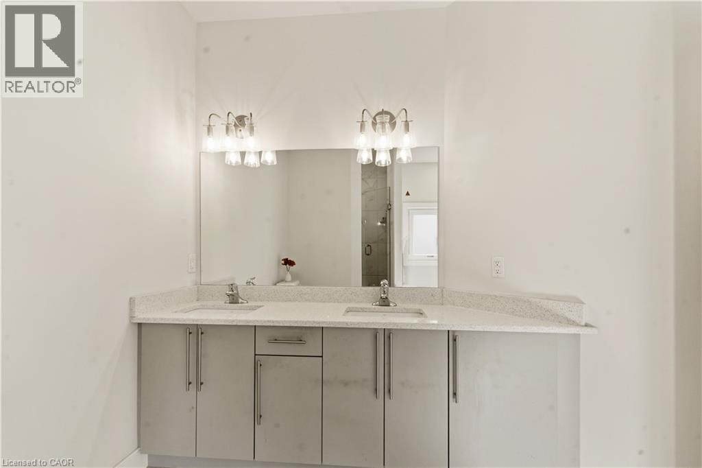 Modern bathroom vanity featuring dual sinks with a light stone countertop, ample cabinet storage with brushed metal hardware, and a large framed mirror illuminated by dual sconce lighting above - 2000 Maddex Way, London, ON - Indoor Photo Showing Bathroom