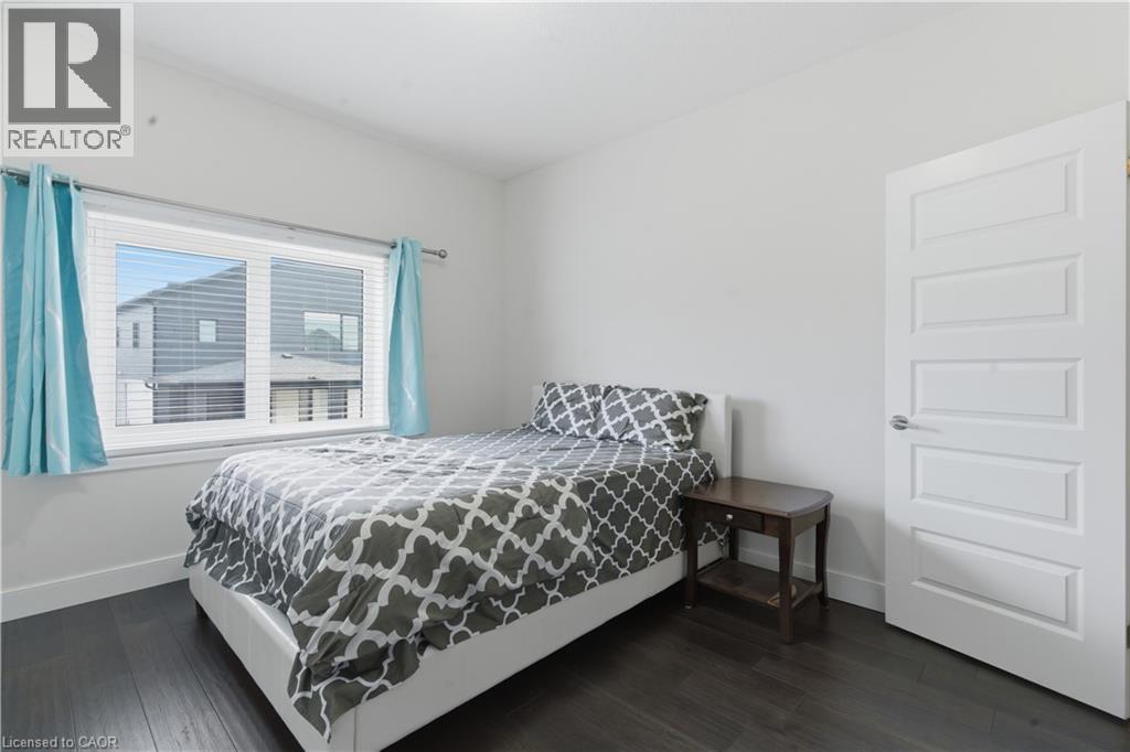 Bright secondary bedroom featuring dark wood-finish flooring, large window with turquoise curtains framing natural light, white six-panel closet door, and wood nightstand - 2000 Maddex Way, London, ON - Indoor Photo Showing Bedroom