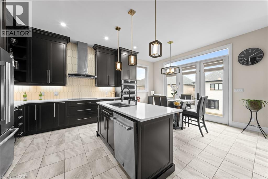 Modern kitchen featuring dark cabinetry, stainless steel appliances, a center island with sink, stylish pendant lighting, and light-toned tile flooring - 130 Pondcliffe Drive, Kitchener, ON - Indoor Photo Showing Kitchen With Double Sink With Upgraded Kitchen