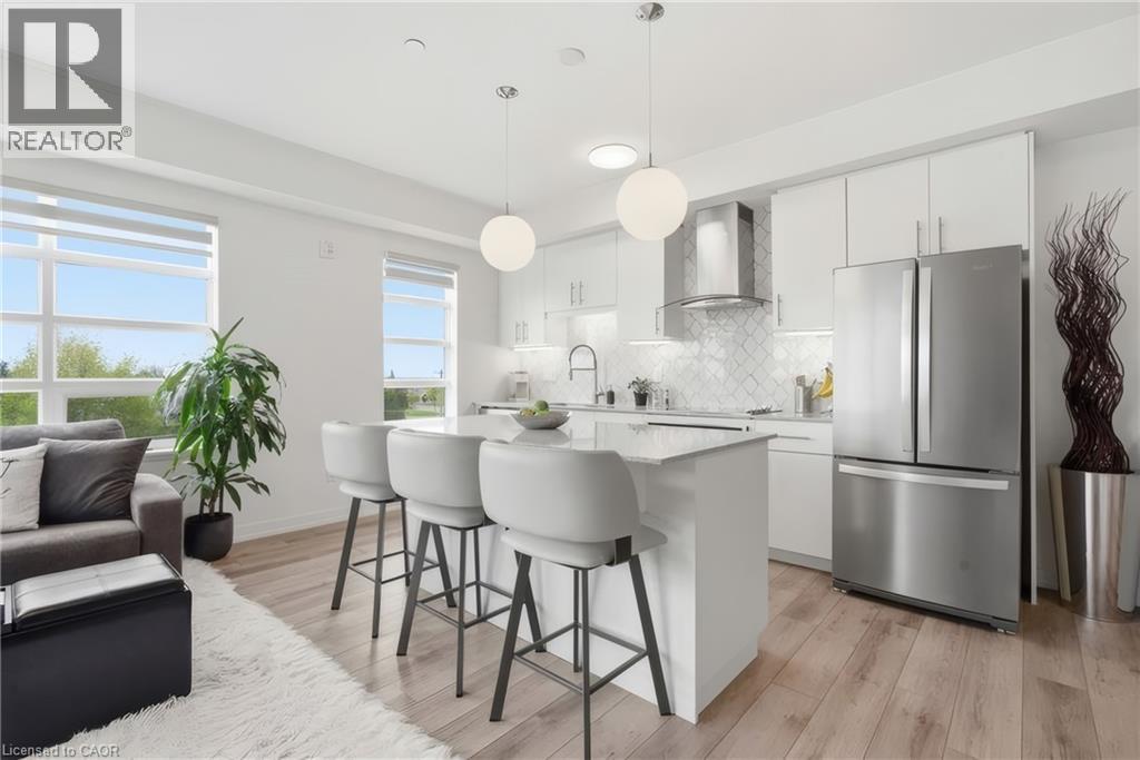 Open-concept living area featuring wood-finish flooring, a kitchen island with seating, pendant lighting, stainless steel appliances, and large windows - 251 Northfield Drive E Unit# 312, Waterloo, ON