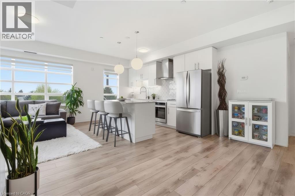 Open-concept living space featuring wood-finish flooring, a kitchen island with seating, stainless steel appliances, white cabinetry, and large windows - 251 Northfield Drive E Unit# 312, Waterloo, ON