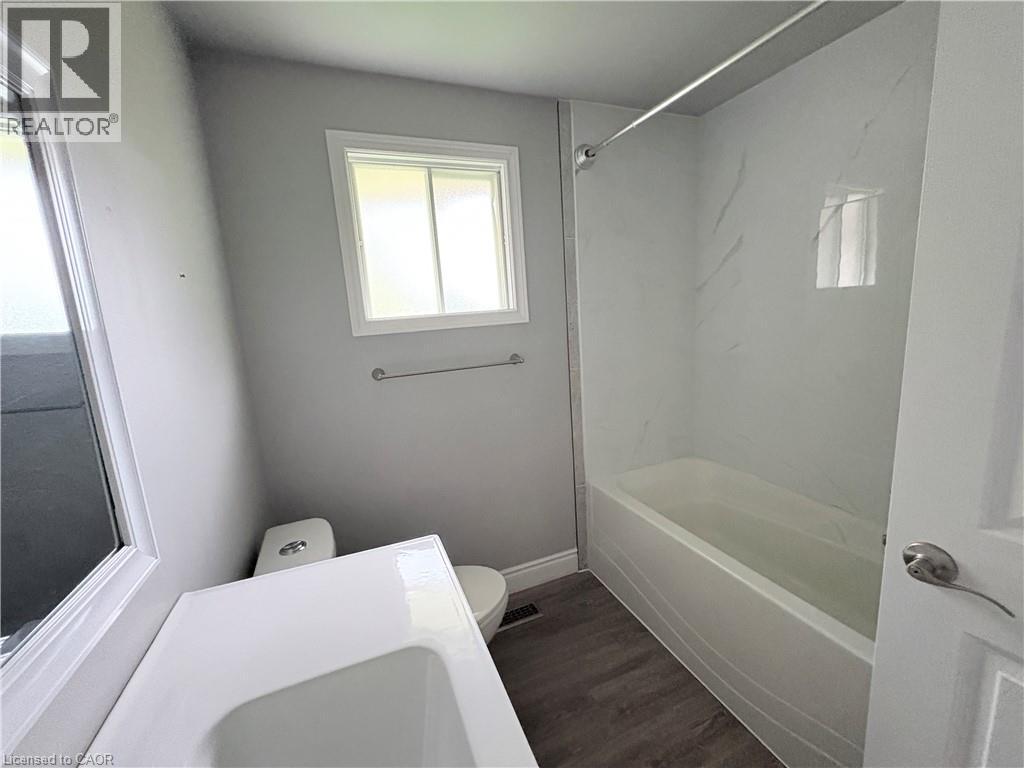 Modern bathroom featuring a white vanity with integrated sink, white bathtub with a marble-patterned surround, wood-finish flooring, and a white framed window - 216 Brunswick Crescent, London, ON