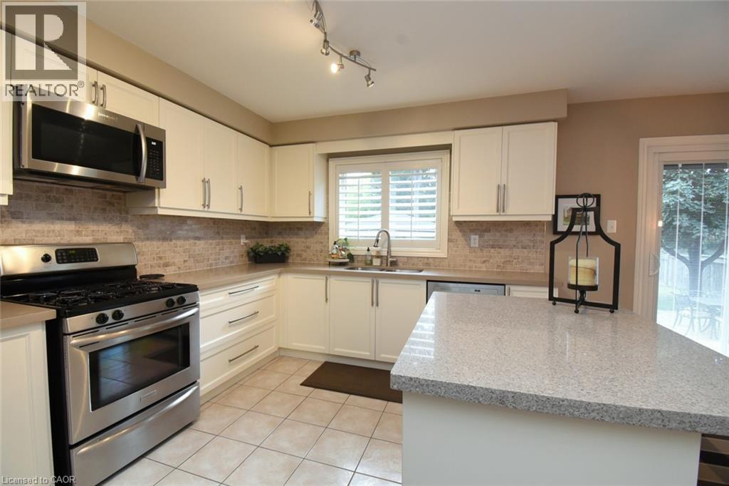 White cabinetry with brushed nickel hardware, stainless steel appliances, tiled backsplash, light-colored countertops, and an eat-at island - 470 Amanda Crescent, Burlington, ON - Indoor Photo Showing Kitchen With Double Sink