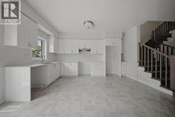 Open-concept space featuring L-shaped white cabinetry, light-toned tile flooring, and a staircase with wood-finish railings - 