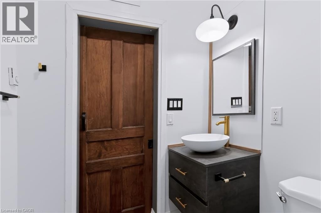 Wood-panel door, modern vanity with vessel sink, gold-finish faucet, wall-mounted mirror with integrated lighting, and contemporary wall sconce - 442 King William Street, Hamilton, ON - Indoor Photo Showing Bathroom