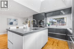 Modern kitchen featuring a skylight, light wood-finish flooring, white and gray cabinetry, integrated stainless steel appliances, and a central island with a sink - 