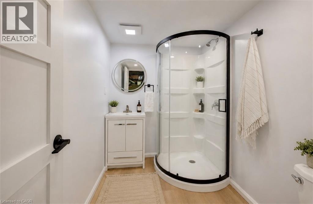 Curved glass shower enclosure with matte black trim, white vanity with integrated sink, circular mirror, and wood-finish flooring - 244 Royal Street, Waterloo, ON - Indoor Photo Showing Bathroom
