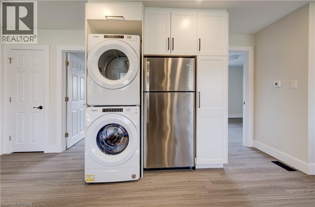 Integrated laundry appliances featuring a stacked washer and dryer, stainless steel refrigerator, white shaker-style cabinetry with matte black hardware, and wood-finish flooring - 361 Prospect Avenue, Kitchener, ON - Indoor Photo Showing Laundry Room