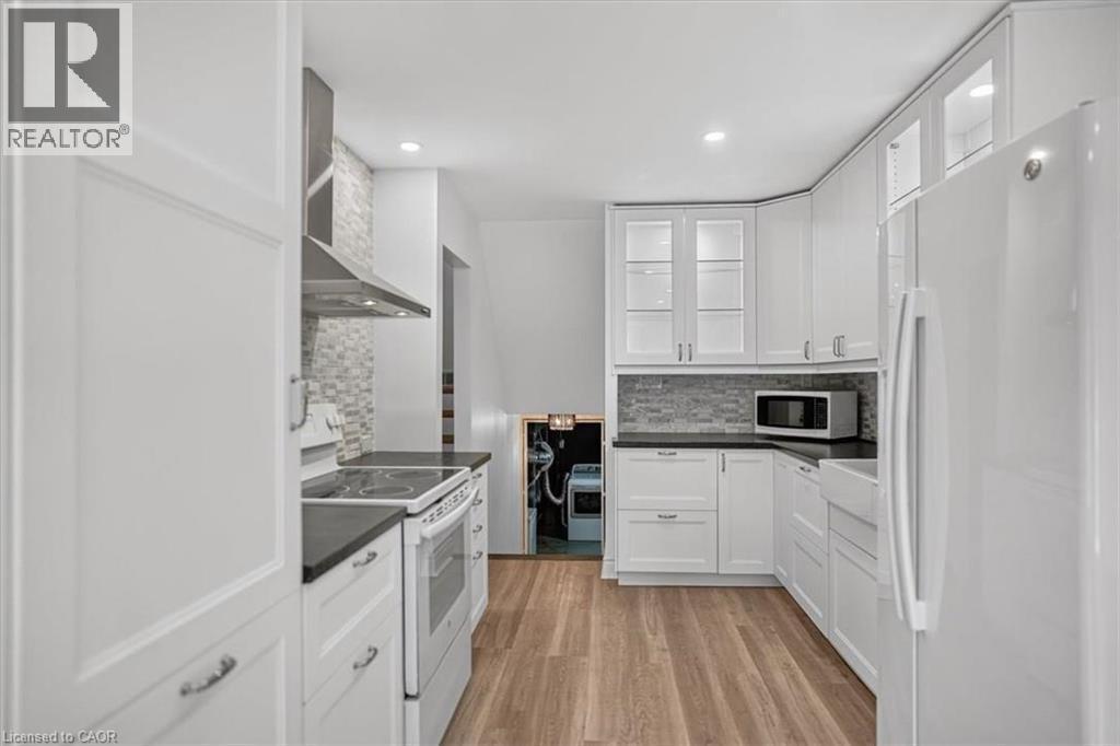 White cabinetry with glass-front uppers, dark countertops, a tile backsplash, stainless steel range hood, and wood-finish flooring - 27 Gretna Court, Hamilton, ON - Indoor Photo Showing Kitchen