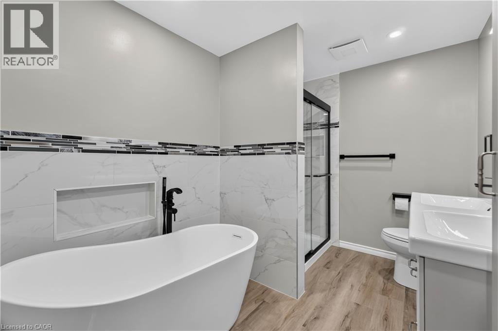 Freestanding soaking tub with a floor-mounted faucet, walk-in shower with glass enclosure, single vanity with integrated sink, wood-finish flooring, and wall-mounted tile accents - 66 Summercrest Drive Unit# 3, Hamilton, ON - Indoor Photo Showing Bathroom