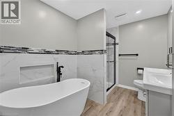 Freestanding soaking tub with a floor-mounted faucet, walk-in shower with glass enclosure, single vanity with integrated sink, wood-finish flooring, and wall-mounted tile accents -