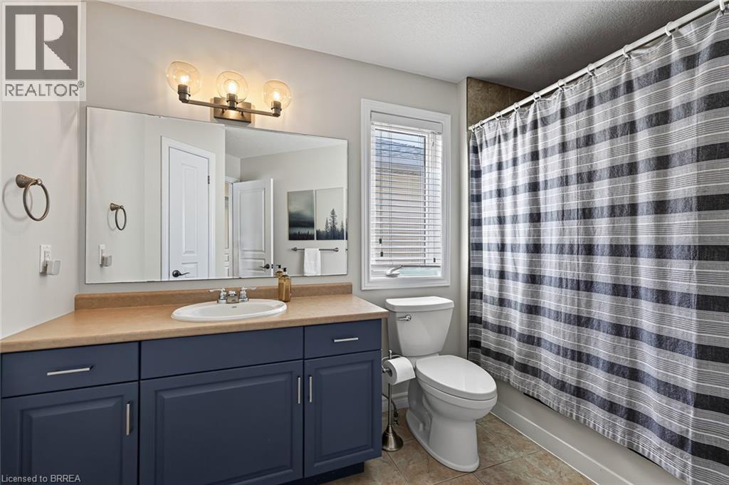 Vanity with a built-in sink and dark blue cabinetry, bronze-finish wall-mounted light fixture, large wall mirror, toilet, and tile flooring - 783 Frontenac Crescent, Woodstock, ON - Indoor Photo Showing Bathroom