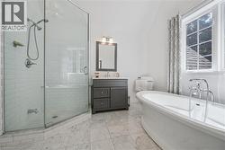 Freestanding soaking tub, glass-enclosed shower with subway tile surround, gray vanity with undermount sink, wall-mounted light fixture, and tiled flooring - 