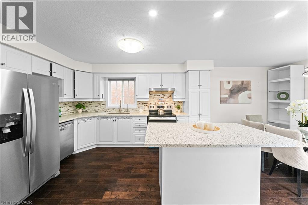Kitchen featuring white cabinetry, a large kitchen island with a light-toned countertop, stainless steel appliances, a tile backsplash, and dark wood-finish flooring - 495 Brigantine Drive, Waterloo, ON