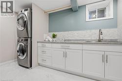 Stacked stainless steel washer and dryer, white shaker-style cabinetry, light-toned countertop, mosaic tile backsplash, and a stainless steel sink with a gooseneck faucet -