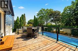 Expansive wood deck with glass panel railing, featuring a rectangular swimming pool, lush perimeter landscaping, and mature trees -