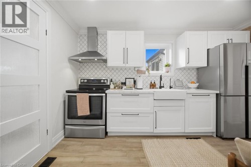 Kitchen featuring white shaker-style cabinetry, stainless steel appliances, a white farmhouse sink, wood-finish flooring, and a patterned tile backsplash - 146 Campbell Avenue, Hamilton, ON 