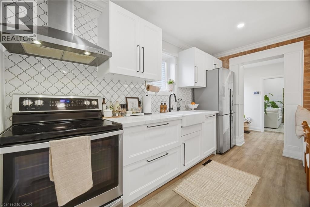 White shaker-style cabinetry with black hardware, stainless steel appliances, geometric tile backsplash, farmhouse-style sink, and wood-finish flooring - 146 Campbell Avenue, Hamilton, ON
