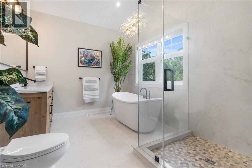 Freestanding soaking tub, glass-enclosed shower with pebble-tile floor, wood-finish vanity with stone countertop, and contemporary chandelier - 293 North Shore Boulevard E, Burlington, ON