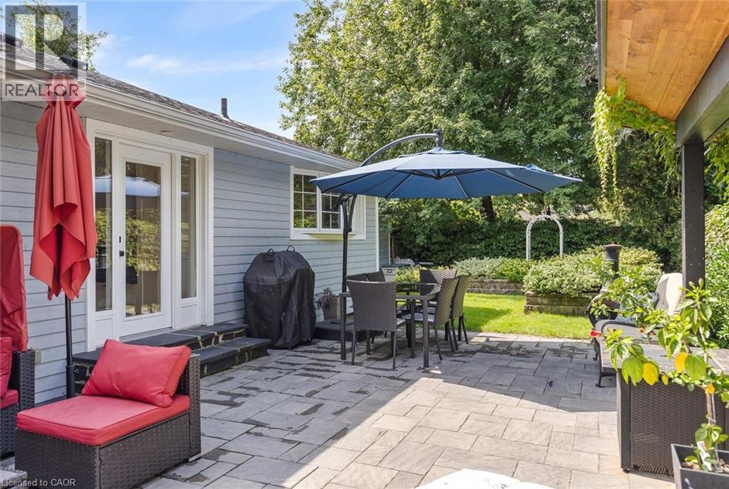 Expansive paver patio with a covered outdoor living area, direct access to the interior via glass panel doors, and a landscaped garden with mature greenery - 293 North Shore Boulevard E, Burlington, ON