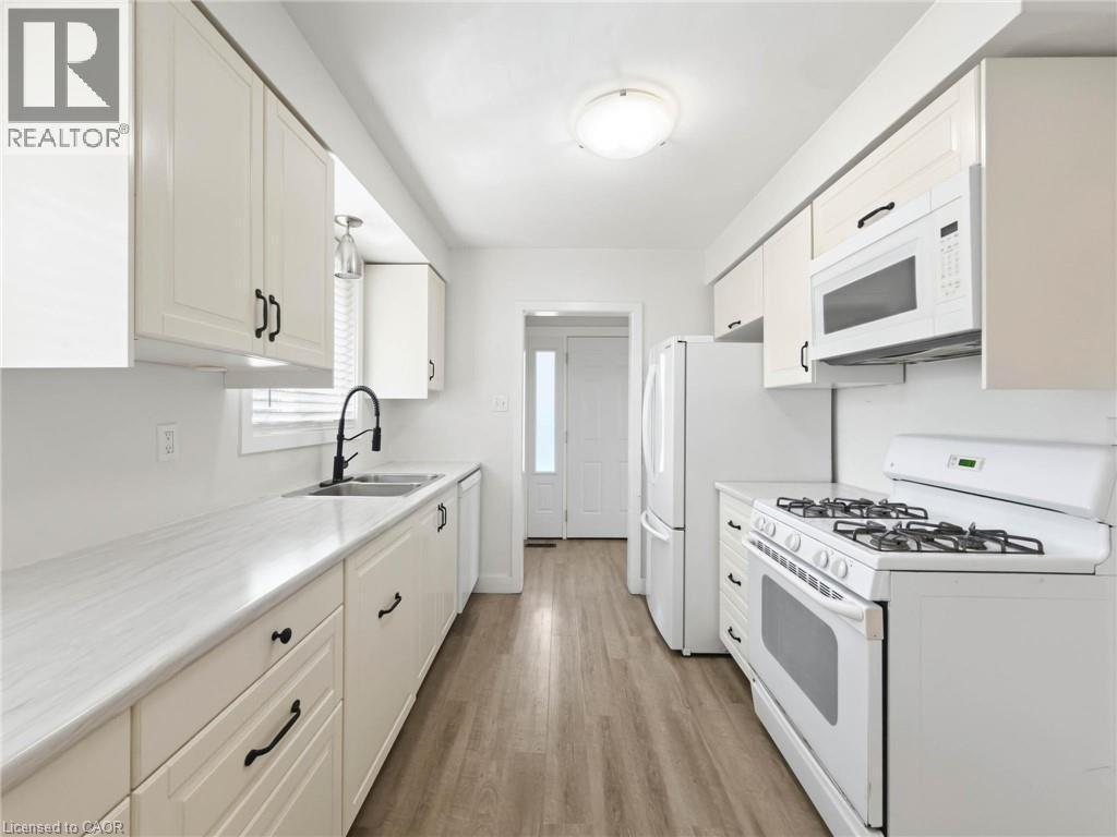 Galley kitchen featuring wood-finish flooring, white cabinetry, light countertops, a stainless steel sink with a matte black faucet, and white appliances - 220 Cairn Street, London, ON