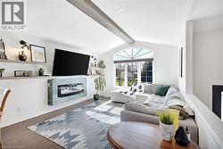 Living area featuring a vaulted ceiling with exposed wood beam, arched window, wood flooring, built-in electric fireplace with tiled surround, and recessed lighting - 