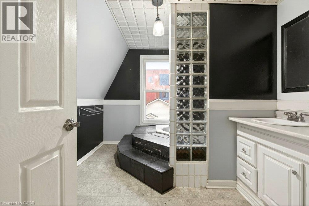 Angled ceiling with pressed tin ceiling tile, window, built-in tub with dark surround, glass block partition, and white vanity with sink - 23 Frederick Avenue, Hamilton, ON