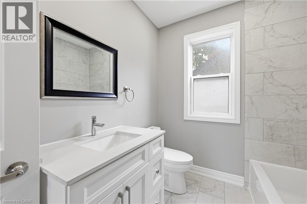 White vanity with a single basin sink and chrome faucet, framed mirror, and tiled walls surrounding the tub - 98 Duke Street Unit# 2Nd, Hamilton, ON