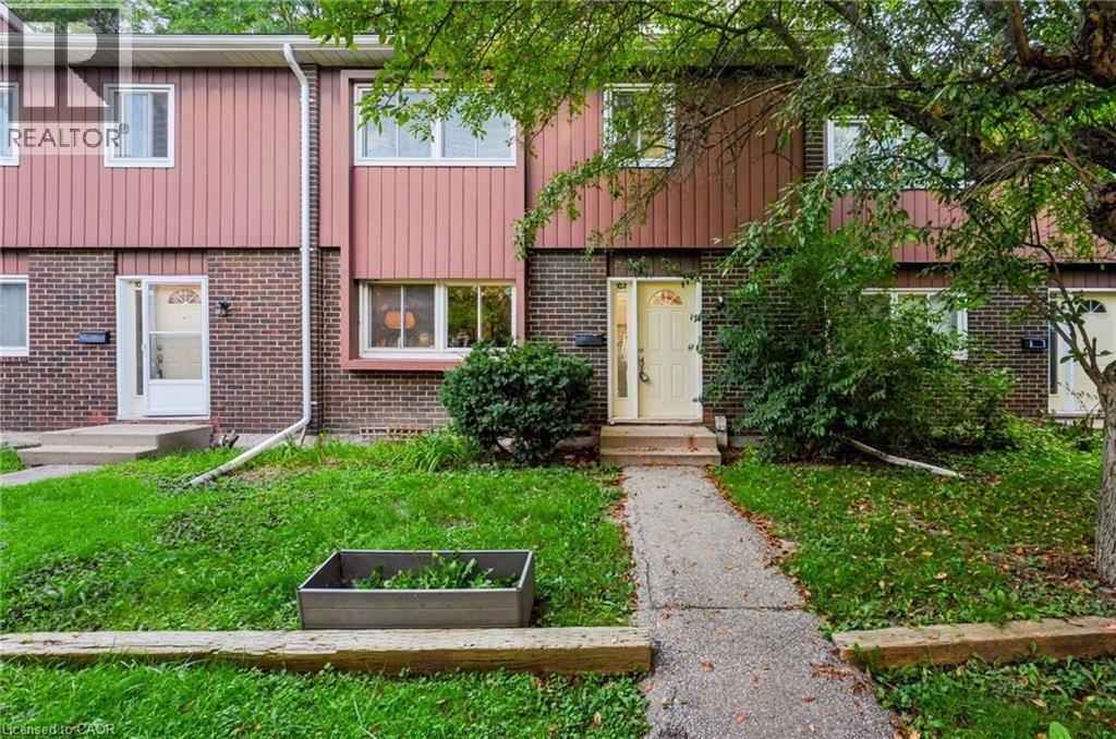 Welcoming townhome entry featuring a brick and metal-panel exterior, mature tree-lined landscaping, and a manicured front lawn with established plantings - 121 University Avenue E Unit# 102, Waterloo, ON