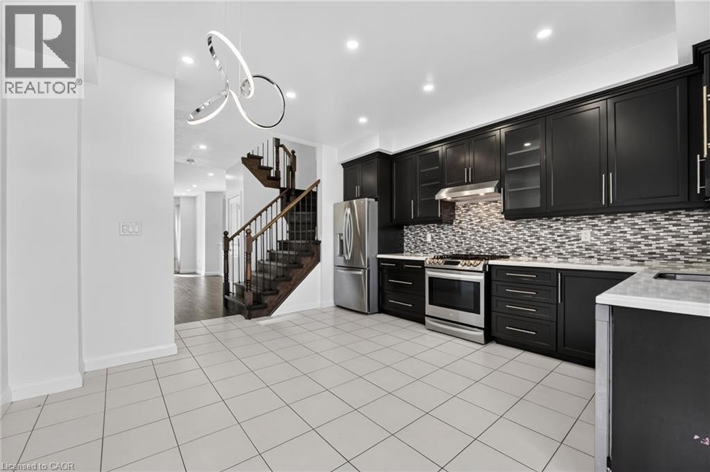 Kitchen featuring dark cabinetry, stainless steel appliances, mosaic tile backsplash, and white tile flooring - 1169 E Garner Road E Unit# 66, Ancaster, ON
