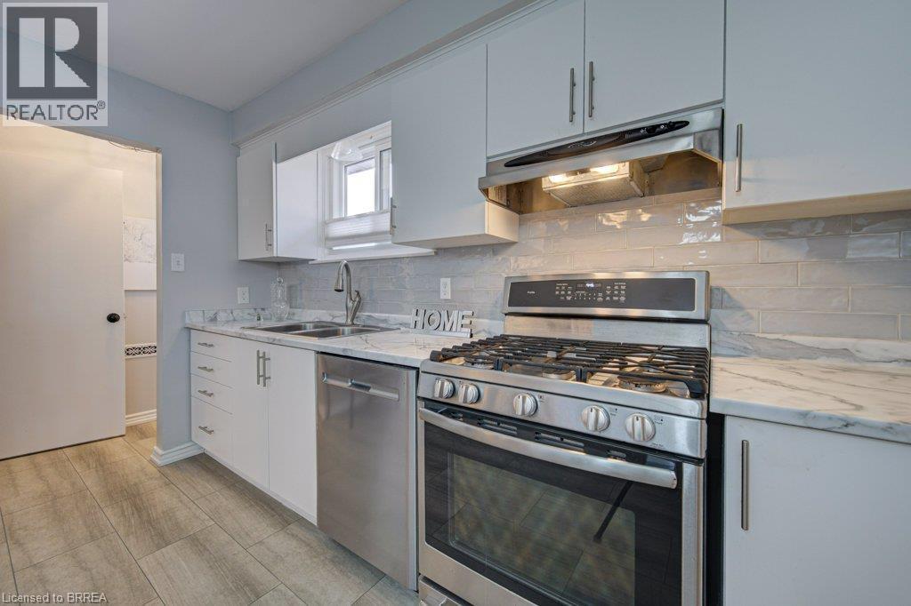 Kitchen featuring stainless steel appliances, wood-finish flooring, white cabinetry with silver hardware, light-toned backsplash, and a double basin sink - 3158 Palmer Drive, Burlington, ON