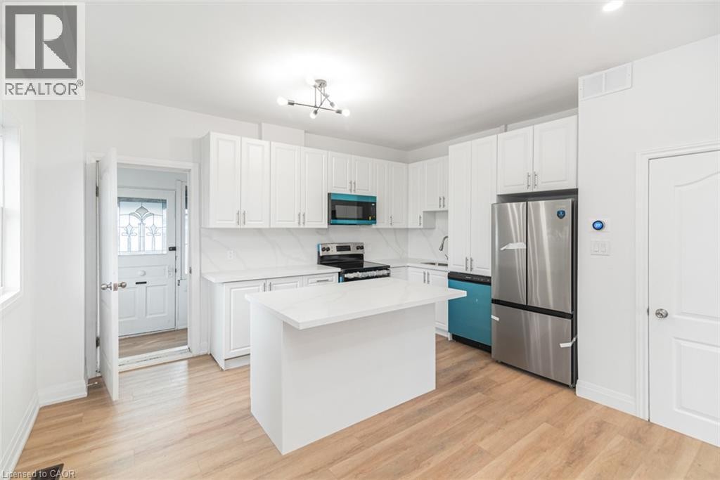 Kitchen featuring wood-finish flooring, white cabinetry, a central island with a solid surface countertop, stainless steel appliances, and an attached ceiling light fixture - 90 Belview Avenue, Hamilton, ON