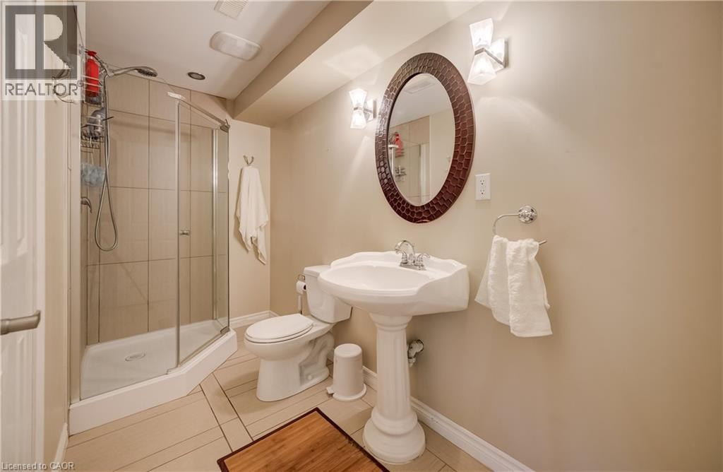 Elegant bathroom featuring a pedestal sink with chrome fixtures, curved glass-enclosed shower stall, ceramic tile surround, wood-finish flooring, and an oval framed mirror with decorative detailing - 573 Robert Ferrie Drive, Kitchener, ON